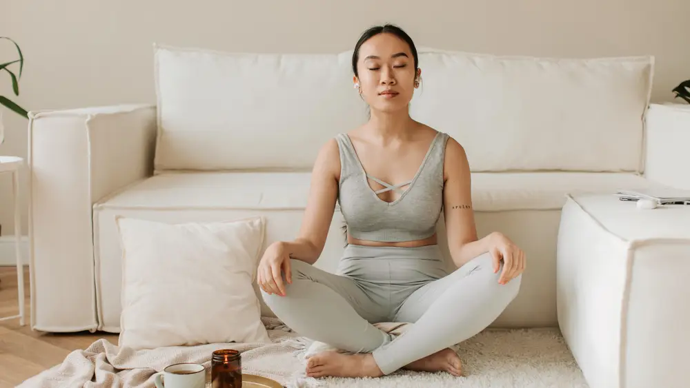 Mulher sentada em meditação, de olhos fechados, num quarto claro